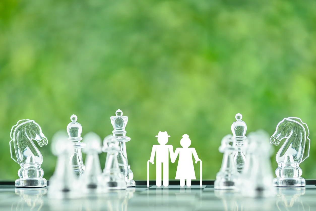 Glass chess pieces on a chessboard with an elderly couple holding hands and using walking canes